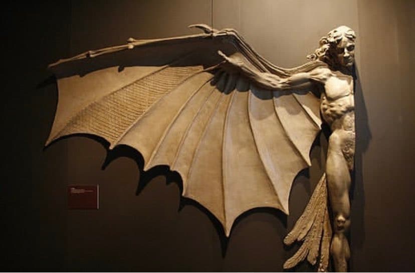 A muscular nude man with long curling hair stands against a dark wall, one arm extended into a huge ribbed, bat-like wing — Artificial Wings, after Leonardo da Vinci