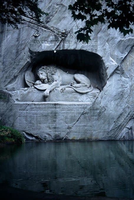 A dying lion with a broken spear in its side lies in a cave carved into a gray sandstone cliff above a still pond — Bertel Thorvaldsen and Lukas Ahorn, The Lion of Lucerne, 1820–21