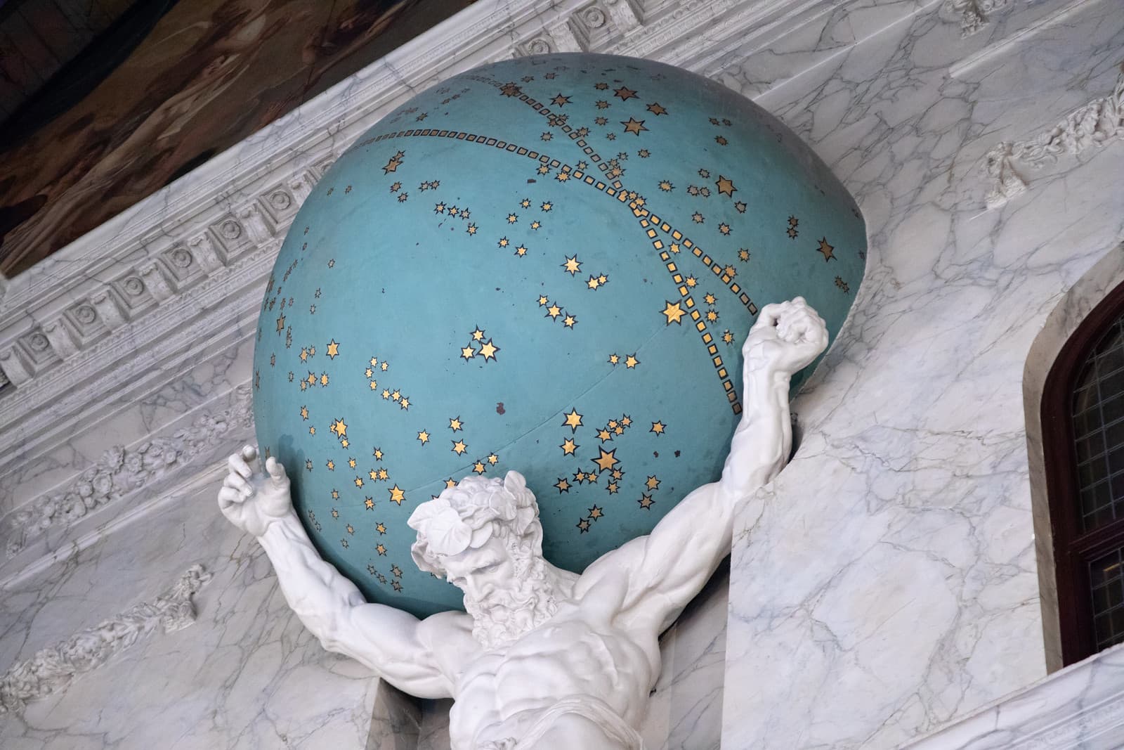 A bearded white figure of Atlas strains with raised arms beneath a blue celestial globe scattered with gold stars, set into a marble wall — Artus Quellinus, Atlas, Royal Palace Amsterdam, c. 1662–63