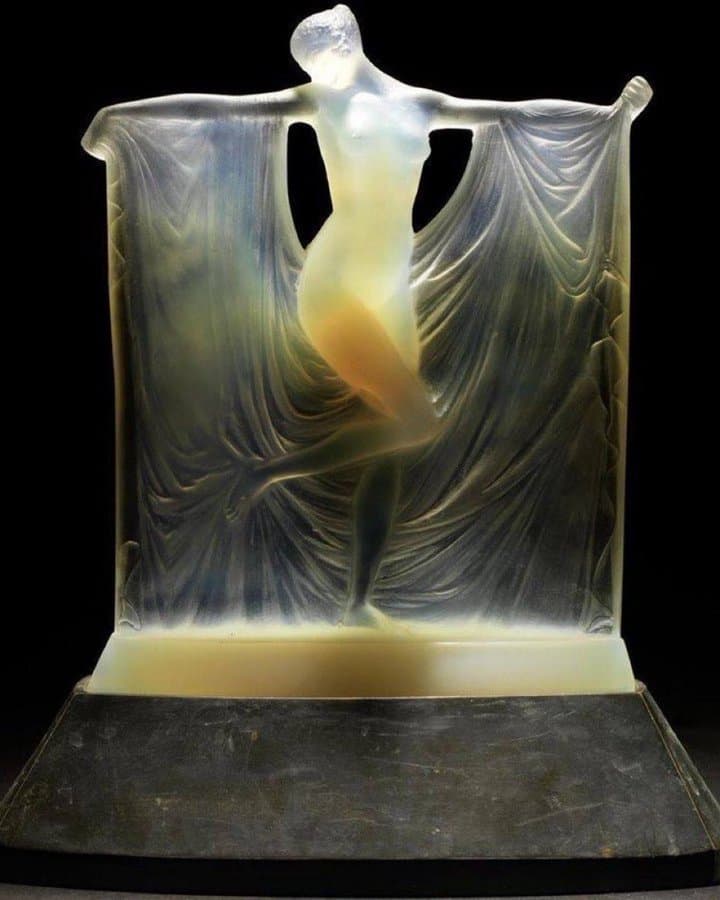 A nude dancer on tiptoe holds a long drapery out from her raised arms, glowing amber in milky opalescent glass on a dark base — René Lalique, Suzanne, 1925