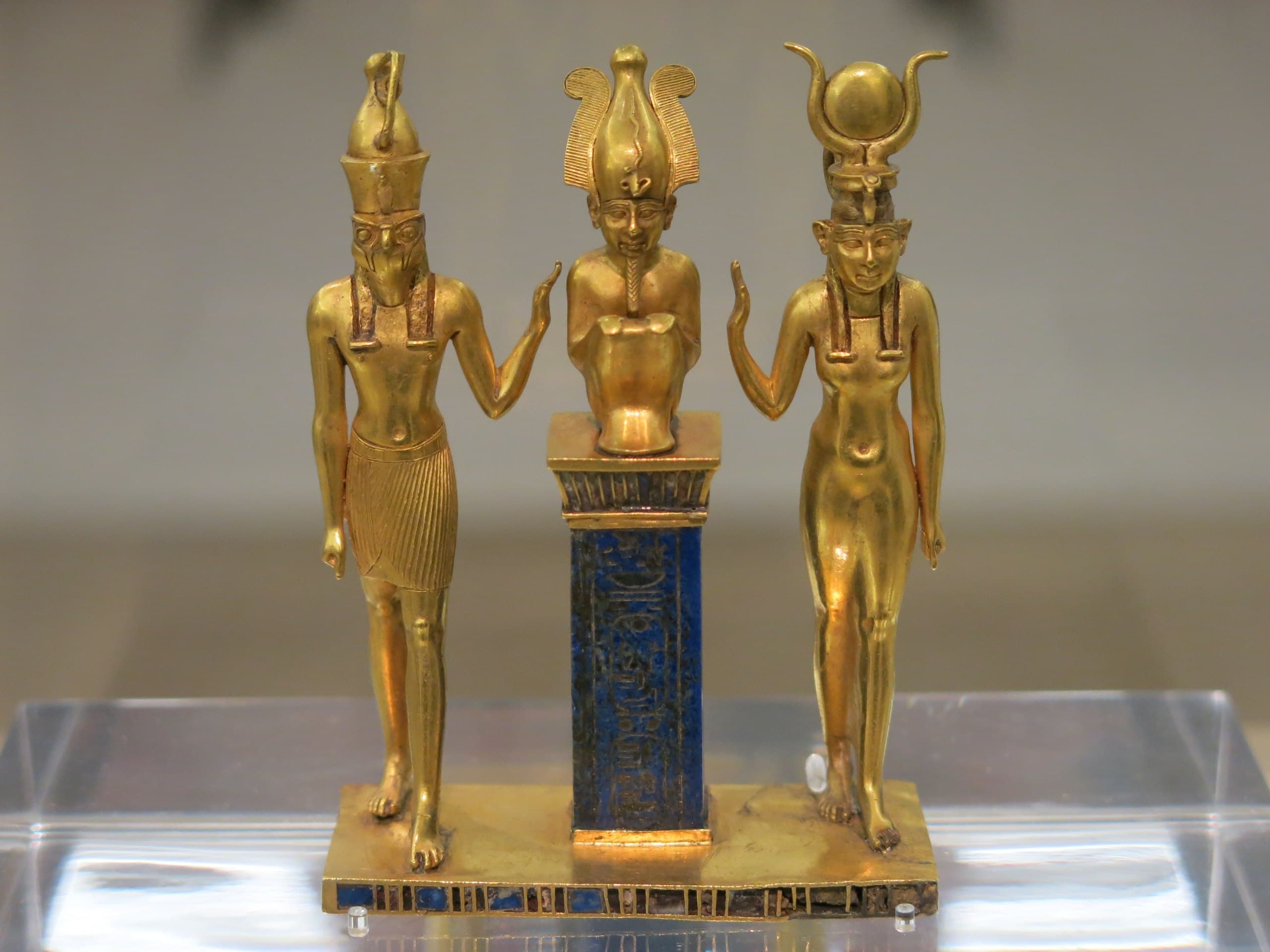 Three small gold figures on a gold base: falcon-headed Horus and Isis with a horned sun disk raise their hands toward Osiris crouching on a blue lapis lazuli pillar — Triad of Osorkon II, Egypt, 9th century BC