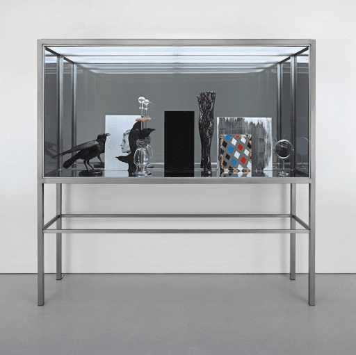 A tall steel-and-glass display case on thin legs holding a stuffed black bird, glass vessels, a black panel, a patterned vase and small paintings — Josephine Meckseper, Afrikan Spir