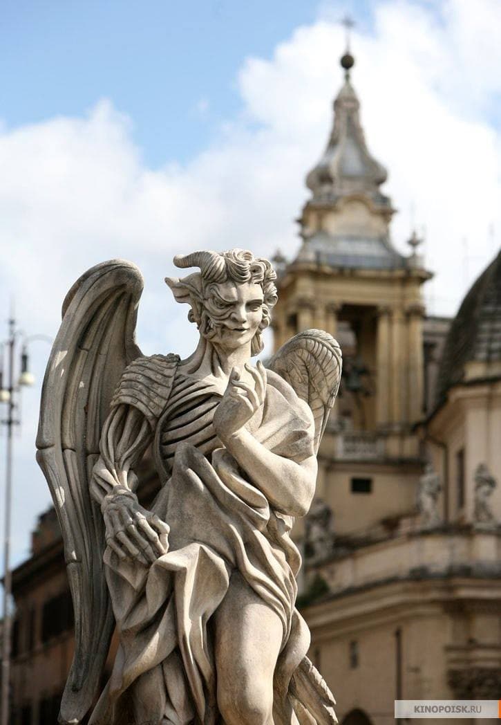 A weathered stone angel with horns, a leering face and an exposed ribcage stands before a church bell tower in a Roman square — prop statue from the film Angels & Demons, 2009