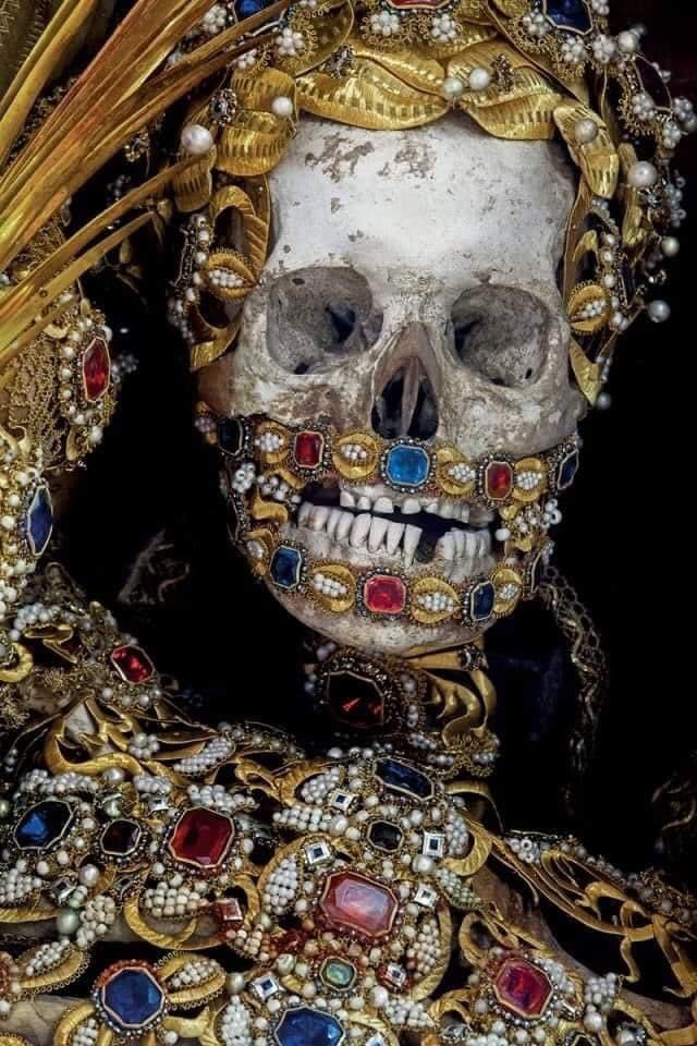 A real human skull framed by a gold-leaf diadem and pearls, its jaw outlined with red and blue jewels, above a breastplate of large faceted stones — Bejeweled Skeleton of Saint Albertus, Burgrain, photographed by Paul Koudounaris