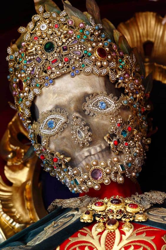 A skull crowned and studded with red, green and blue jewels, with staring glass eyes set in jeweled rosettes, above a red and gold costume — Bejeweled Skeleton of Saint Felix