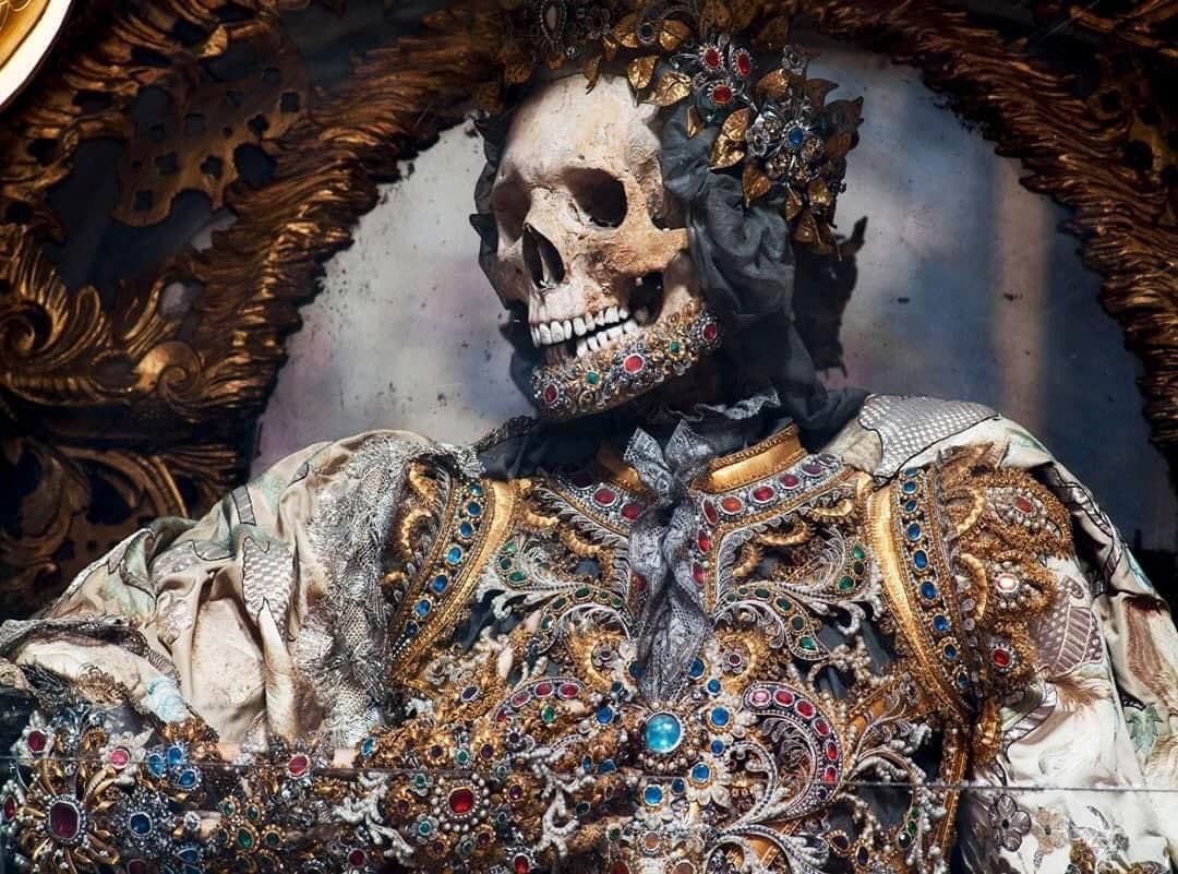 A skull crowned with gilt leaves and jewels, a band of gems along its jaw, above a breastplate of gold scrollwork and colored stones, inside a gilded frame — Bejeweled Skeleton of Saint Gratian, decorated by Adalbart Eder, photographed by Paul Koudounaris