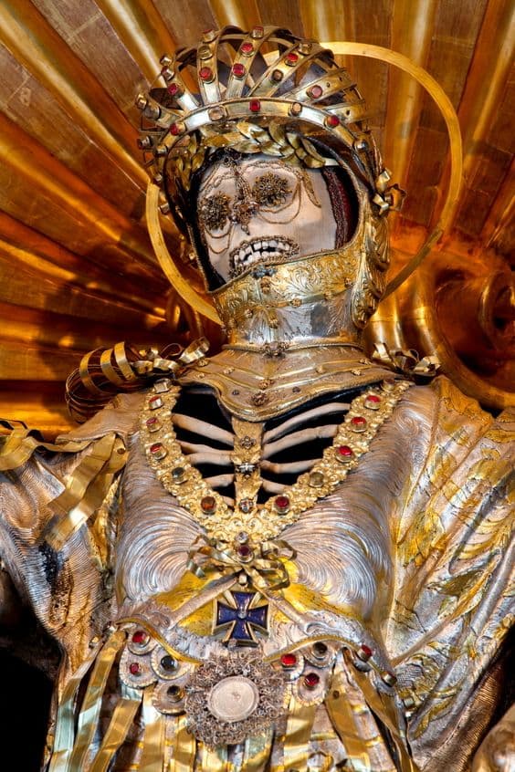 A skeleton in a gilded helmet with raised visor, crowned and haloed, gold filigree in its eye sockets and ribs showing through a silvered breastplate, before a golden sunburst — Bejeweled Skeleton of Saint Maximus, Bürglen, photographed by Paul Koudounaris