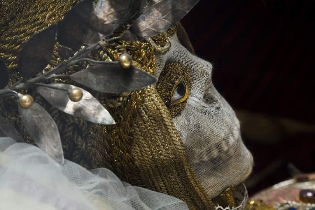 A skull veiled in fine gauze, with a glass eye set in gold embroidery, wears a wreath of silver leaves and braided gold cords — Bejeweled Skeleton of Saint Munditia, St. Peter's Church, Munich