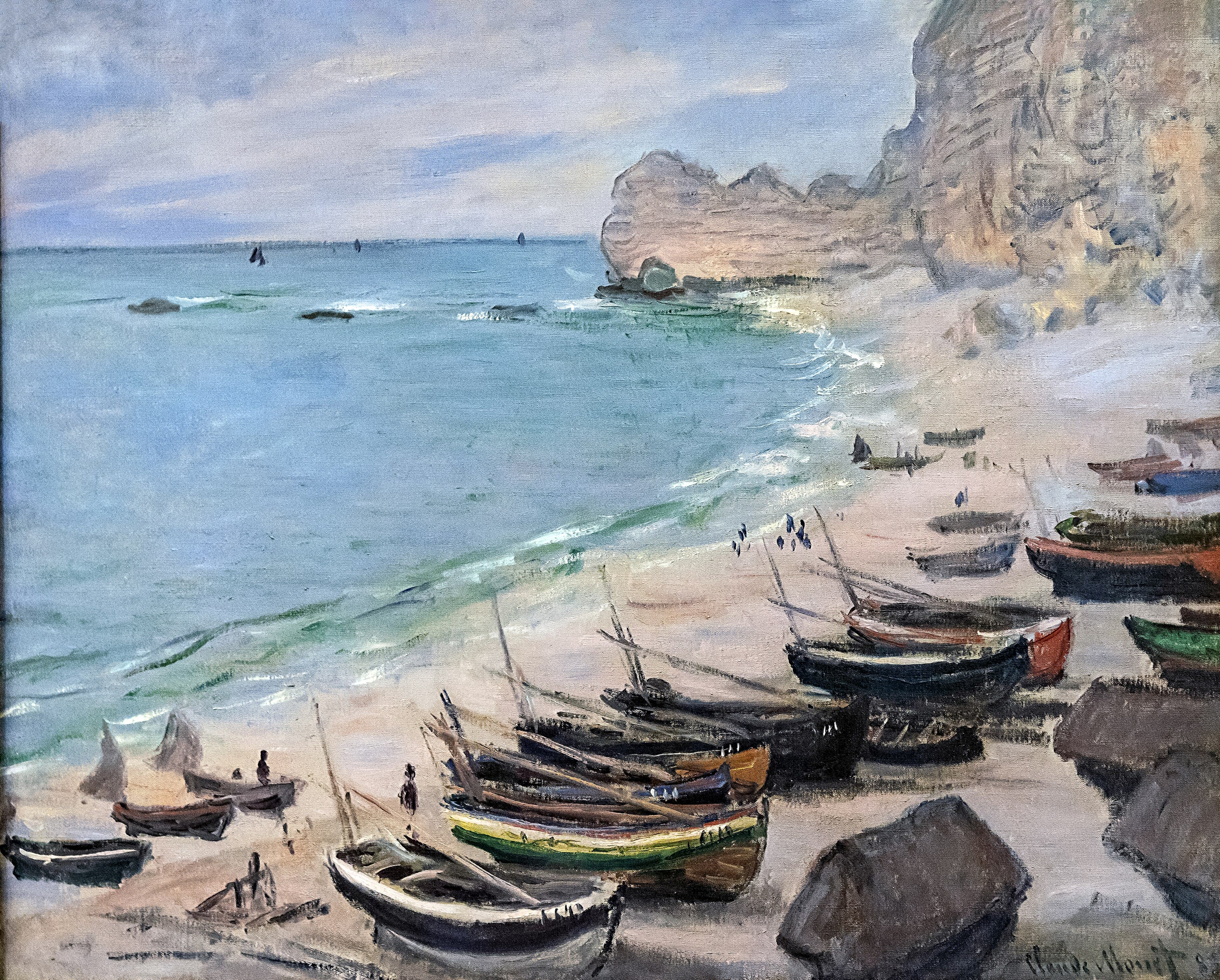 Rows of small fishing boats lie on a curving shingle beach beside a turquoise sea, white chalk cliffs with an arch on the right — Claude Monet, Boats on the Beach at Étretat, 1883