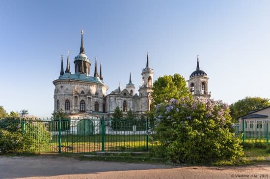 A pale stone church with pointed windows, black spires and a green-blue domed lantern rises behind a green fence and a flowering lilac — Church of the Vladimir Icon of the Mother of God, Bykovo, 1788–89