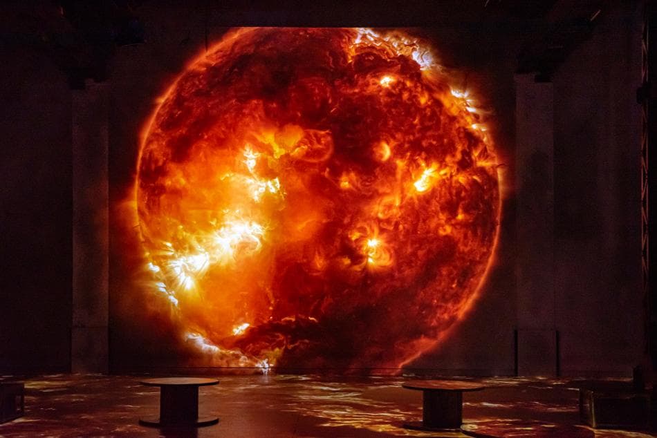 A giant projected image of the blazing orange Sun fills the wall of a dark exhibition hall, with small seats in silhouette in front — Destination Cosmos, 2022