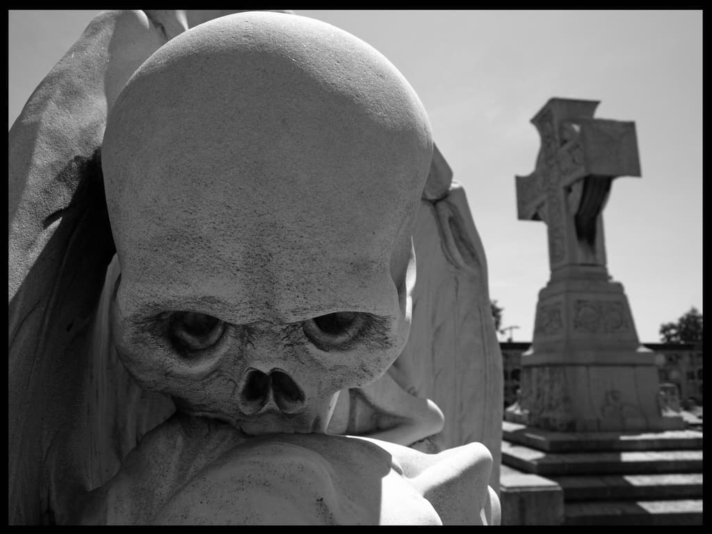 Close view of a marble skull with folded wings pressing its teeth to the head of a young man, with a stone cross in the background — The Kiss of Death (El Petó de la Mort), Poblenou Cemetery, Barcelona, 1930