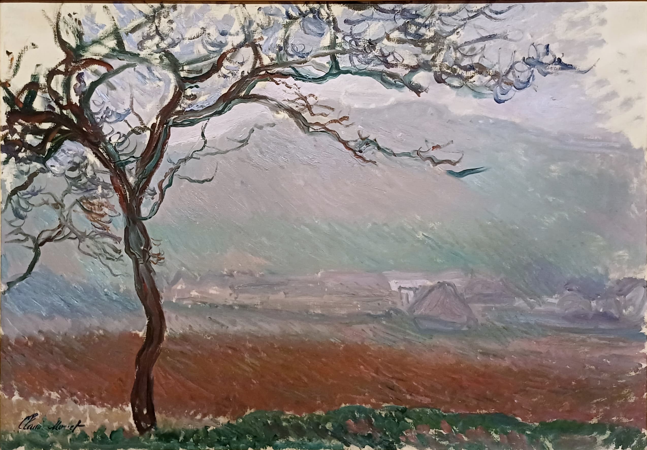 A thin, windswept tree with looping branches stands at the left before a rust-red field and a misty valley with pale houses under a streaked grey-lilac sky — Claude Monet, Giverny Landscape, 1887
