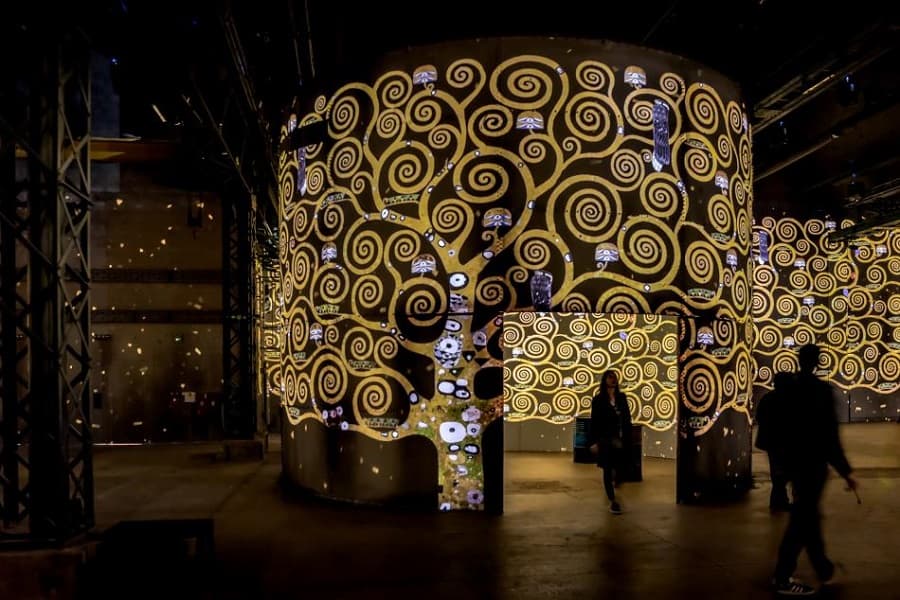 Gold spiraling branches of Klimt's Tree of Life projected onto a tall cylinder in a dark industrial hall, visitors walking past as silhouettes — immersive Gustav Klimt exhibition, 2018