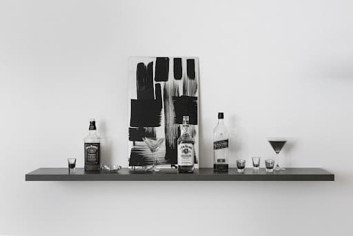 A dark wall shelf holding whiskey bottles, shot glasses and cocktail glasses in front of a black-and-white gestural abstract painting — Josephine Meckseper, Here and Elsewhere