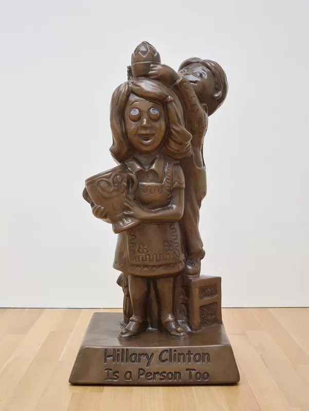 A large brown figurine of a big-eyed girl holding a mug, with a boy on a chair behind her, on a base reading Hillary Clinton Is a Person Too — Jonathan Horowitz, Hillary Clinton Is a Person Too, 2008