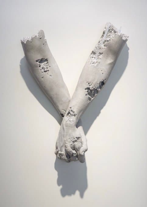 Two grey stone forearms cross on a white wall with hands clasped, their surfaces broken open to show glittering crystals — Daniel Arsham, Holding Hands, 2015