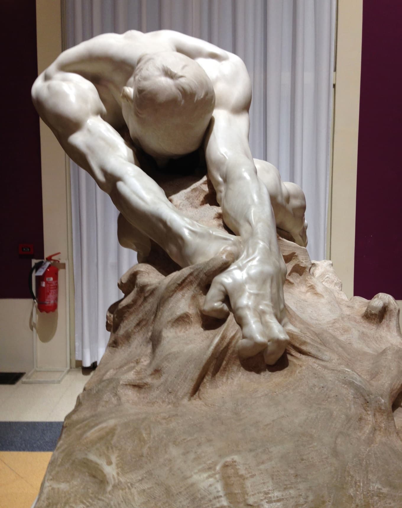 A polished marble nude man crouches with bowed head, his clawed hands gripping a rough, swirling mass of stone beneath him — Gaetano Cellini, Humanity Against Evil (L'umanità contro il male), 1908