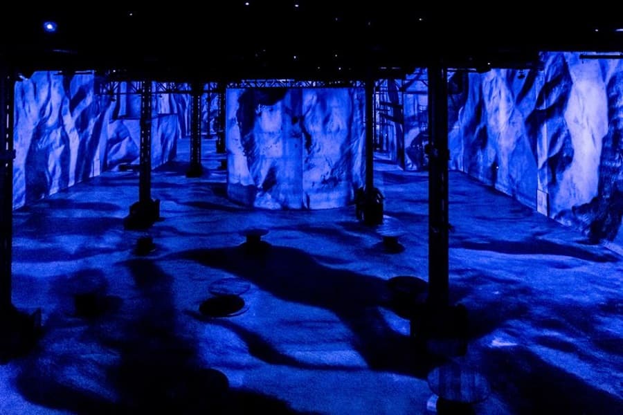 An empty industrial hall with steel columns, its walls, screens and floor flooded with deep blue projected textures like a rocky planetary surface — Yves Klein, l'infini bleu, Atelier des Lumières, Paris, 2020