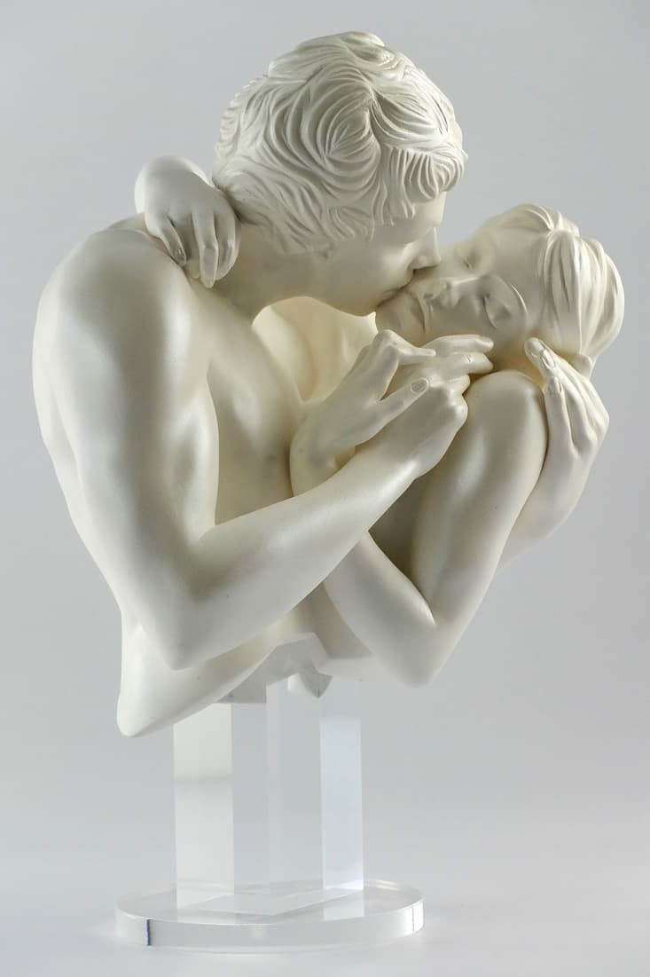 A pearl-white sculpture of a man and woman cut off at the chest, kissing with their arms wrapped around each other, raised on a clear acrylic stand — Yves Pirès, Le Baiser Nacré