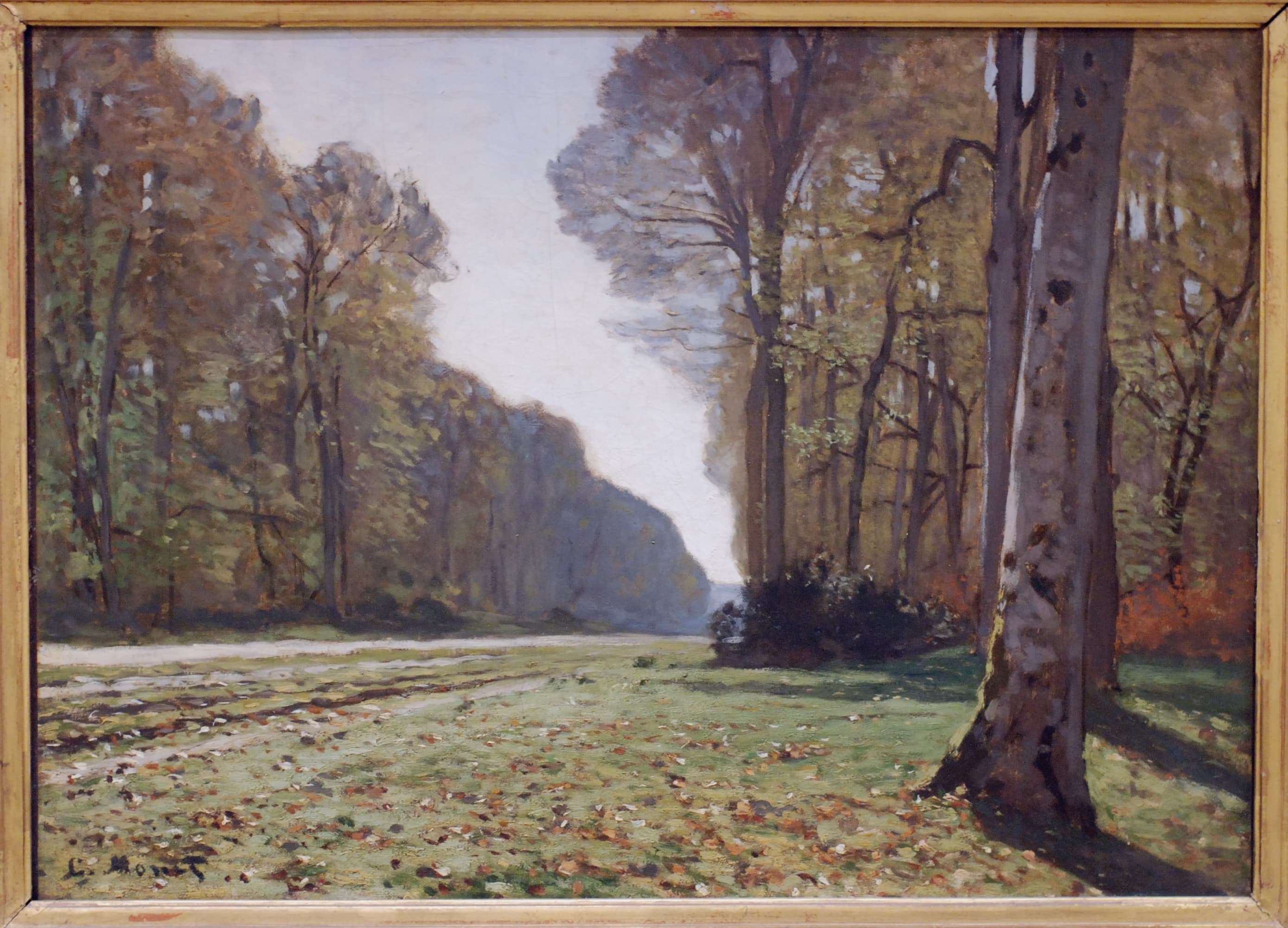 A straight earth road runs between autumn trees into a hazy forest distance, a large trunk on a leaf-covered verge at right — Claude Monet, Le Pavé de Chailly, c. 1865
