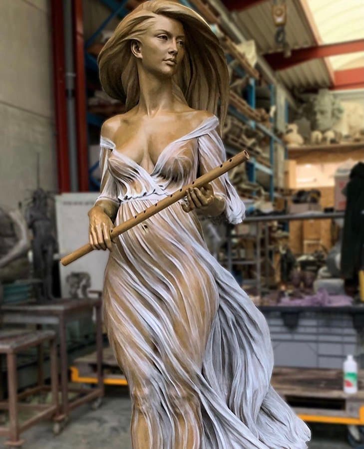 A bronze young woman holds a long flute across her body as wind sweeps her hair and pale, sheer dress behind her, photographed in a foundry — Luo Li Rong, Mélodie, 2020