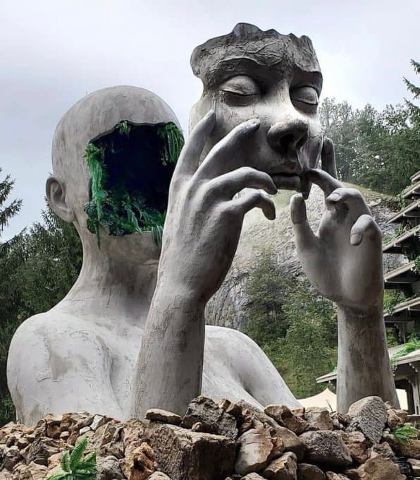 A giant gray figure rising from a pile of rocks holds its serene face out in front of it like a mask, revealing ferns growing inside the hollow head — Daniel Popper, Modem Swamp, 2019