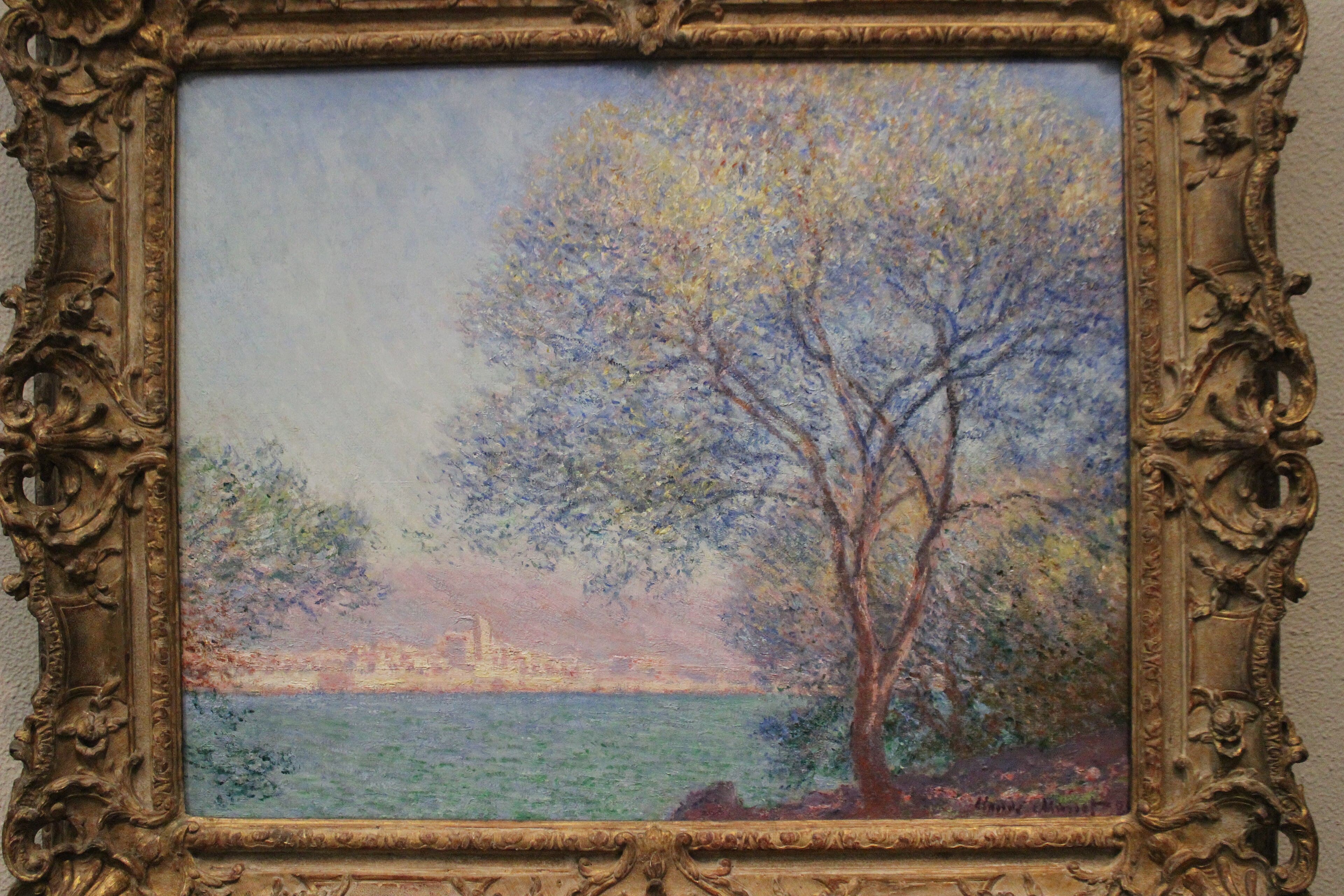 A tall tree with pale rosy branches and lilac foliage stands on the shore, framing a distant pink town across a calm green sea in morning haze — Claude Monet, Morning at Antibes, 1888