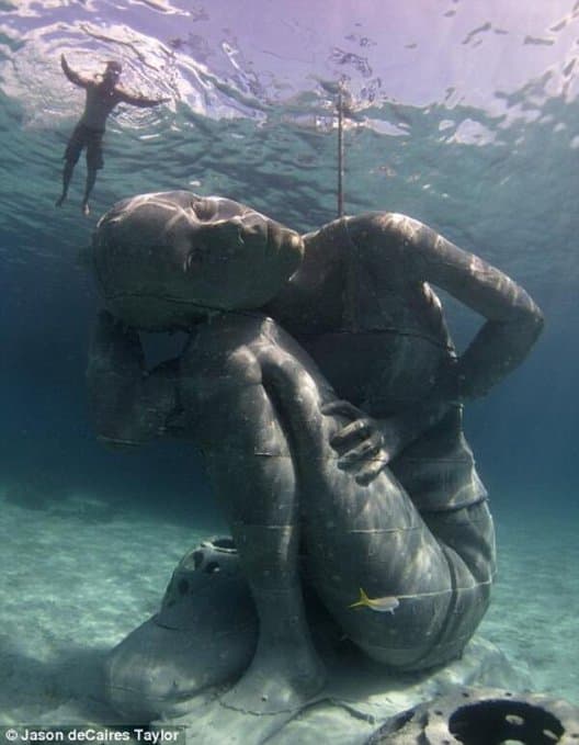 A giant gray sculpture of a crouching girl on the sandy seabed, one arm raised to the surface, a snorkeler floating above — Jason deCaires Taylor, Ocean Atlas, 2014