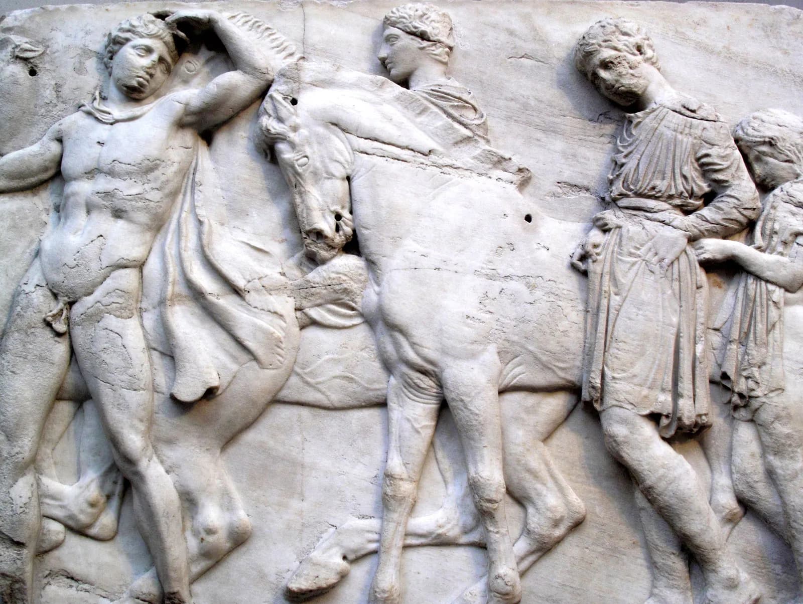 A nude youth with a slipping cloak raises a hand to his head beside a turning horse and a bearded man in a short tunic, carved in shallow marble relief — Parthenon Frieze, c. 443–437 BC
