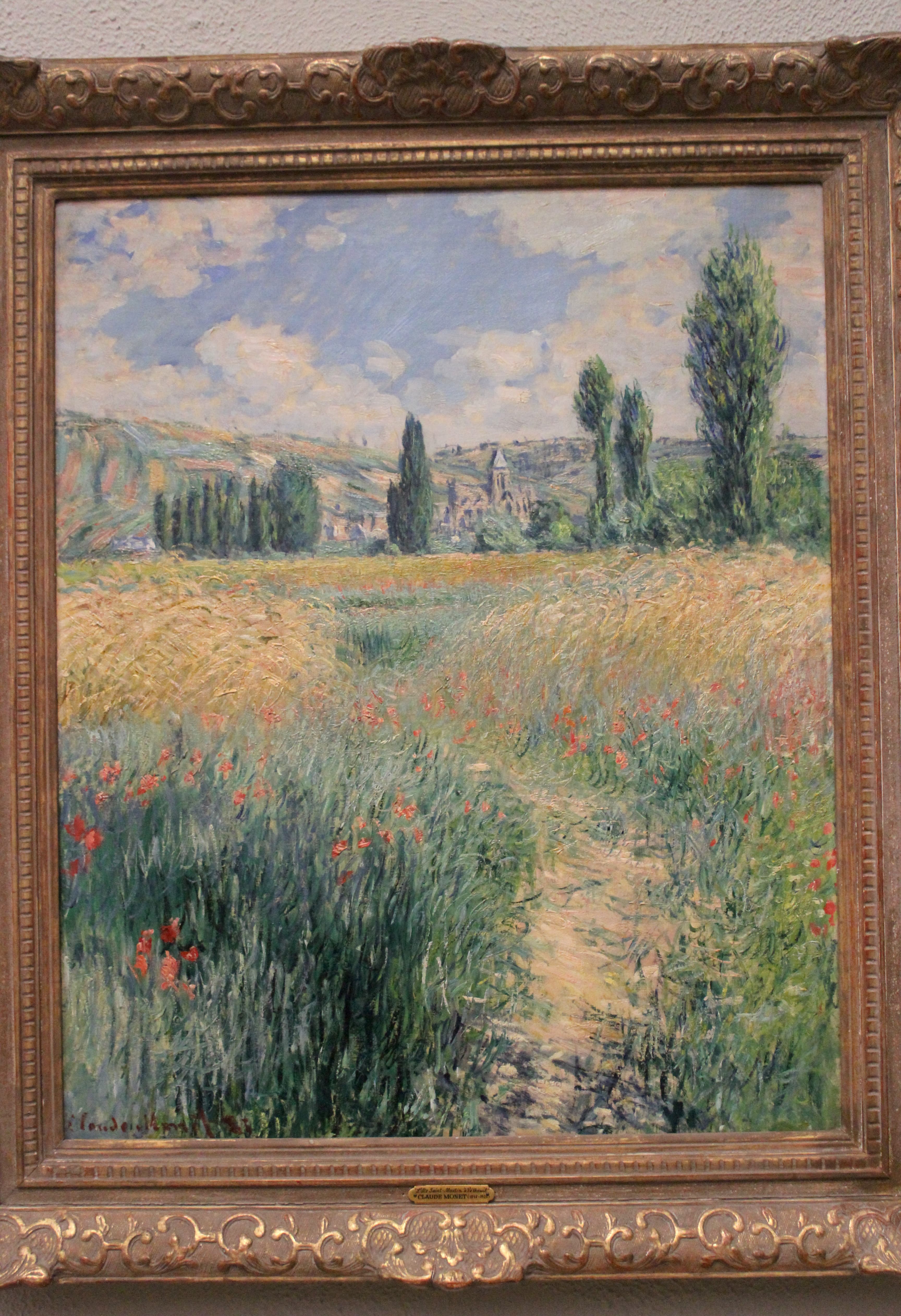 A sandy path winds through green wheat and red poppies toward a village church and tall trees under a blue sky with white clouds — Claude Monet, Path on the Island of Saint Martin, Vétheuil, 1881