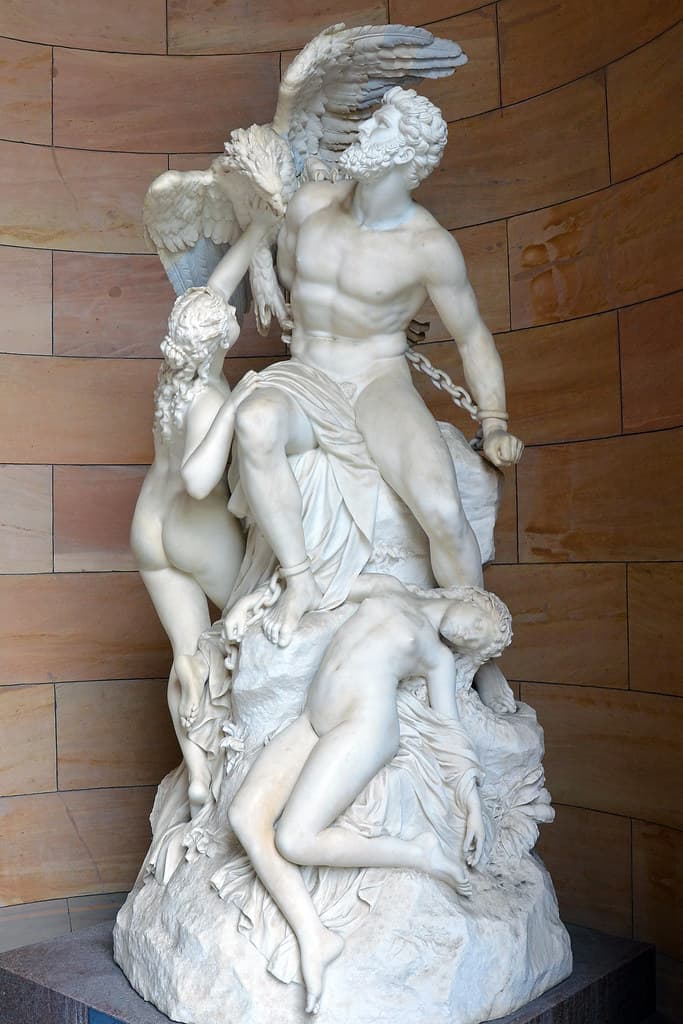 A chained, bearded Prometheus sits on a rock beneath a spread-winged eagle while one nude sea nymph reaches up toward the bird and another lies collapsed at his feet, in white marble — Eduard Müller, Prometheus Bound and the Oceanids, 1879