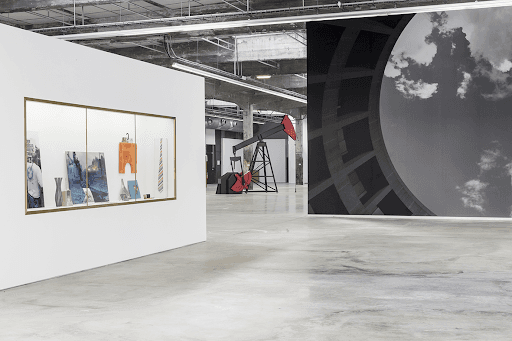 A raw industrial hall holds a gold-framed vitrine of arranged objects on a white wall, a black and red oil pump jack sculpture and a wall-sized black-and-white photograph — Josephine Meckseper, solo exhibition, Nantes, 2019