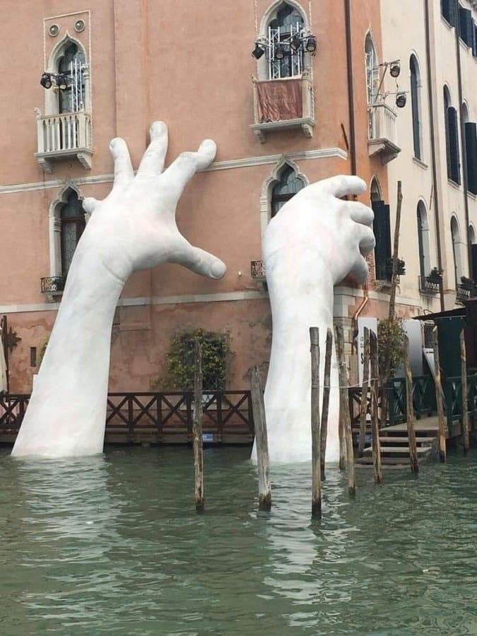 Two giant white hands rise from the Grand Canal and press against the pink façade of a Venetian palazzo — Lorenzo Quinn, Support, 2017