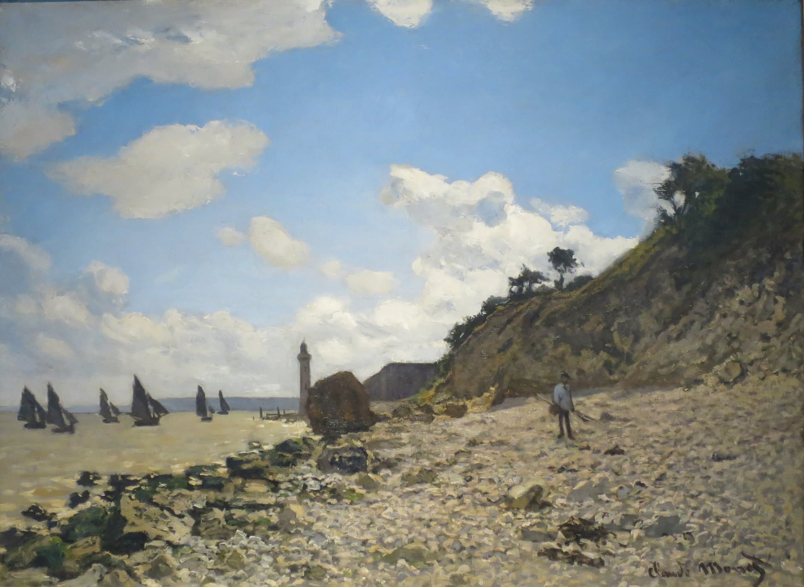 A stony beach below a grassy bluff under heaped white clouds, with dark-sailed fishing boats, a slender lighthouse and a lone fisherman — Claude Monet, The Beach at Honfleur, 1864–66