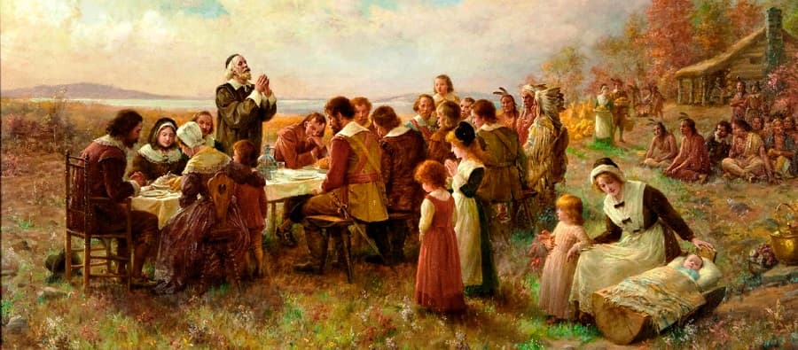 A white-bearded elder prays over a long outdoor table of colonists while Native men in feathered headdresses sit on the grass and a mother tends a baby in a log cradle — Jennie Augusta Brownscombe, The First Thanksgiving at Plymouth, 1914
