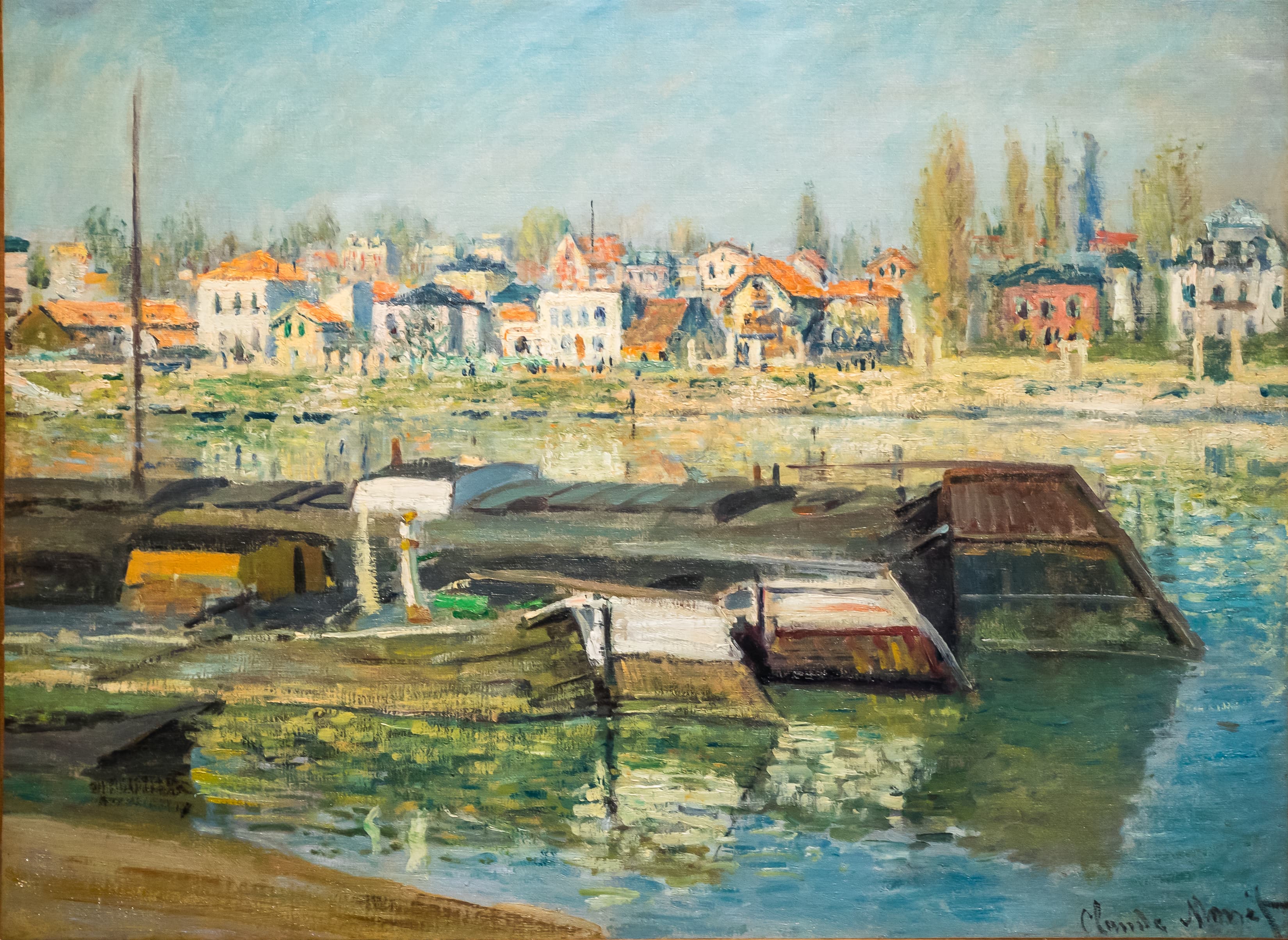 Moored barges and floating wooden sheds on a calm river, with a row of colorful houses and bare trees along the far bank — Claude Monet, The Seine at Asnières, 1873