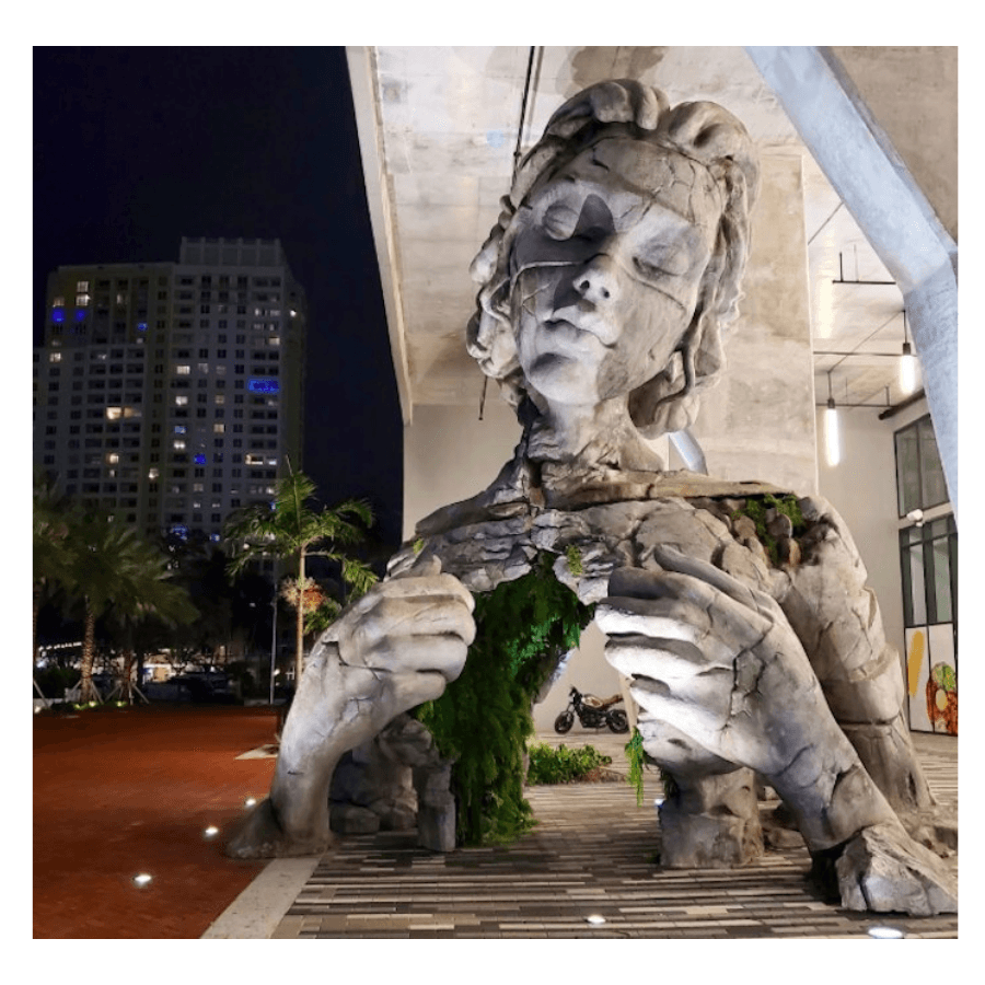 A giant cracked concrete figure with closed eyes pulls open her chest to reveal green ferns, beneath a tower at night — Daniel Popper, Thrive, 2020