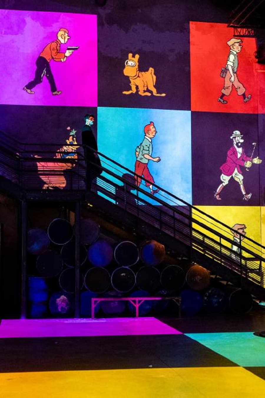 Projected Tintin characters, including Tintin and Snowy, stride across bright colored panels on a dark wall behind an iron staircase — Tintin, l'aventure immersive, 2023