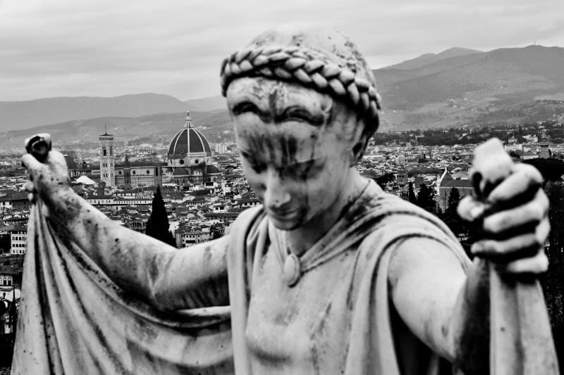A weathered stone statue of a young woman with a braided crown of hair, head bowed, holding her cloak open like wings above the rooftops and cathedral dome of Florence — Tombstone of Silvia Marini nei De Rogati, San Miniato al Monte