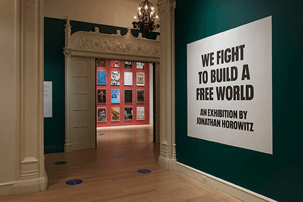 A green gallery wall with a large white sign reading We Fight to Build a Free World, beside a doorway opening onto a red wall of posters — We Fight to Build a Free World, exhibition by Jonathan Horowitz, Jewish Museum, 2020