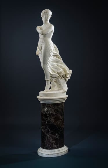 A white marble statue of a young woman with crossed arms stepping forward as wind sweeps her skirt back, on a dark marble pedestal — Thomas Ridgeway Gould, The West Wind, modeled 1870