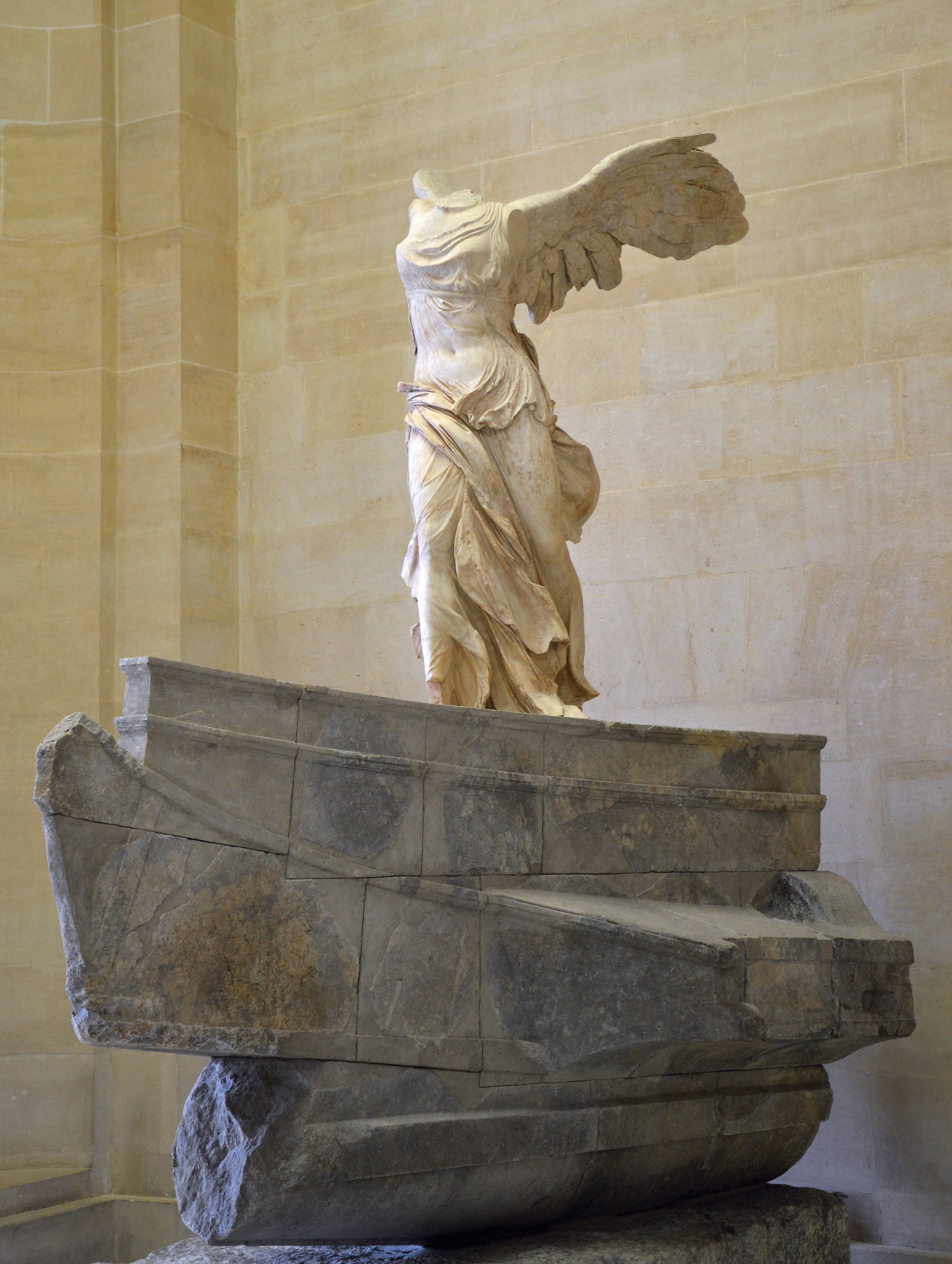 A headless winged marble goddess in wind-swept drapery strides forward on the prow of a stone ship at the top of a grand staircase — Winged Victory of Samothrace, c. 200–190 BC