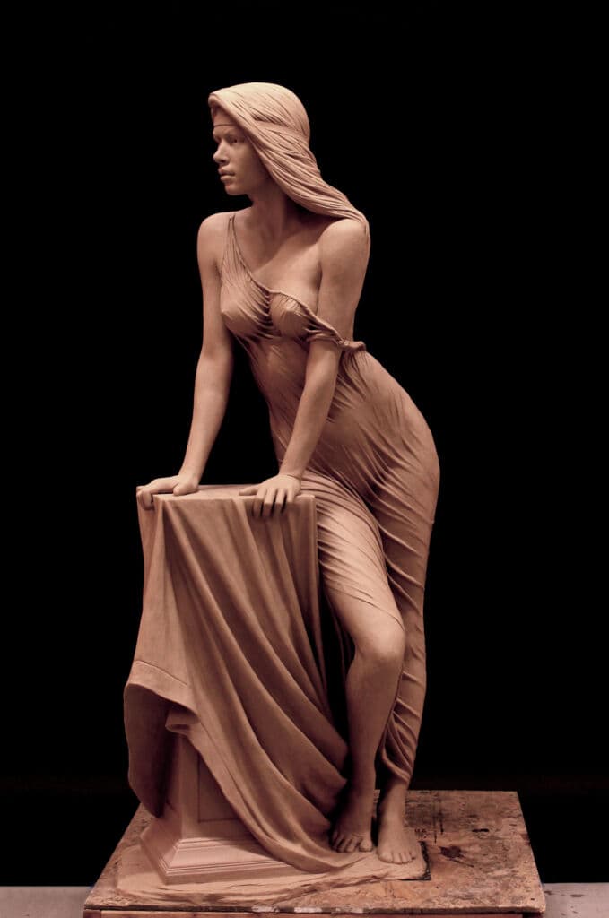 A young woman in clinging, rippling drapery leans on a cloth-covered pedestal and turns her head sharply to one side — Benjamin Victor, Zuleikha, clay, c. 2022